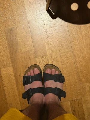 Svarta sandaler med spännen  - Svarta sandaler med två breda remmar och justerbara metallspännen. Sulan är platt och har en naturlig brun färg. Perfekta för varma dagar och enkel att matcha med olika outfits.