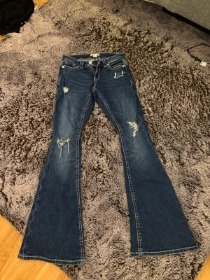 Blå bootcut jeans med slitningar - Snygga blå bootcut jeans med slitna detaljer och broderade bakfickor. Jeansen har klassisk femficksdesign och markerade sömmar. Perfekta för dig som gillar en retroinspirerad look med lite edge.