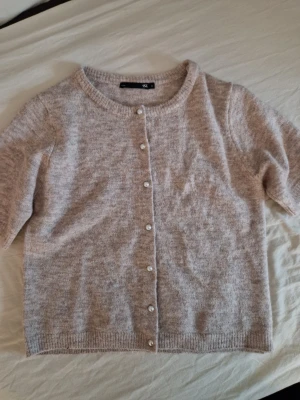Beige kofta - Mysig beige kofta från lager 157 med rund halsringning och pärlknappar framtill. Koftan har korta ärmar och är stickad i ett mjukt material. Enkel och stilren design som är lätt att matcha. Oanvänd, men Lapparna saknas. Hund finns I hemmet.