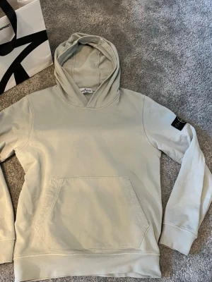  Stone Island hoodie - Säljer en stilren ljusgrå hoodie från Stone Island med klassisk huva och stor magficka. Ikoniska Stone Island-märket sitter på vänster ärm. Hoodien är gjord i mjuk bomull och har ribbade muddar vid ärmslut och nederkant.