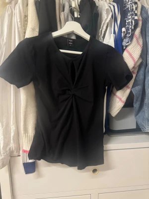Svart topp med twistdetalj  - Svart kortärmad topp från Polen med twistad detalj och liten öppning framtill vid halsen. Toppens passform är normal och materialet känns mjukt och stretchigt, perfekt för en trendig look.