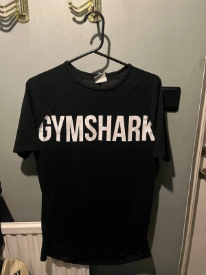 Svart Gymshark t-shirt - Svart t-shirt från Gymshark med stor vit logga över bröstet. Klassisk rund halsringning och korta ärmar. Perfekt för träning eller en sportig look. Tillverkad i ett lätt och stretchigt material som andas. Muscle fit