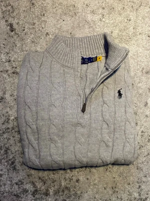 Grå Ralph Lauren quarter-zip - En elegant kabelstickad quarter-zip i grått med svart logga. Levererar behaglig upplevelse på, och en stilren look samtidigt. Storleken är M fast passar som en mindre S enligt mig, skicket är mycket bra eftersom den är näst intill oanvänd. Hör gärna av dig vid funderingar så löser vi det! 😁