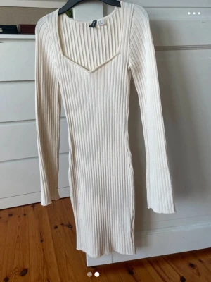 Vit ribbad stickad klänning - Säljer en vit ribbad stickad klänning med lång ärm och v-ringad hals. Klänningen är kort och har en figurnära passform som framhäver siluetten. Perfekt för dig som gillar en enkel men snygg look.