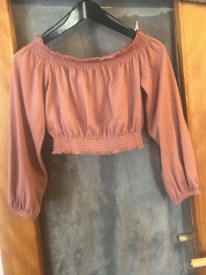 Rosa croppad offshoulder topp - Trendig croppad offshoulder topp i dammigt rosa med smockdetaljer vid halsringning och midja. Lätt puffiga trekvartsärmar ger en romantisk vibe. Perfekt till höga jeans eller kjol.