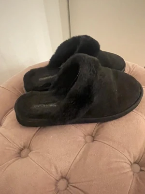 Svarta fluffiga tofflor från Even&Odd - Mysiga svarta tofflor från Even&Odd med fluffig pälsimitation och rund tå. Perfekta att glida runt i hemma, med mjuk insida och platt sula. Klassisk slip-in modell som är enkel att ta på och av.