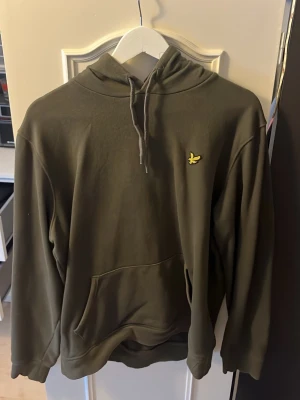 Olivgrön hoodie från Lyle & Scott - Snygg olivgrön hoodie från Lyle & Scott med klassisk gul logga på bröstet. Hoodien har huva med dragsko och en stor magficka framtill. Perfekt för en avslappnad och stilren look. Tillverkad i mjukt material för extra komfort.