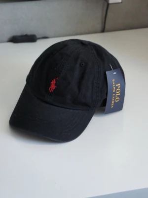 Svart keps från Polo Ralph Lauren - Snygg svart keps från Polo Ralph Lauren med klassisk böjd skärm och röd broderad logga framtill. Justerbar rem baktill för perfekt passform. Tillverkad i bomull för en skön och avslappnad look. OBS - liten i storlek