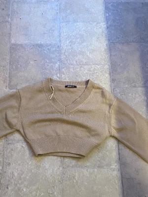 Beige cropped stickad tröja Gina Tricot - Trendig beige cropped stickad tröja från Gina Tricot med v-ringning och bred ribbad mudd nedtill. Perfekt att styla med höga jeans eller kjol. Mjuk och skön kvalitet, långärmad och enkel att matcha.
