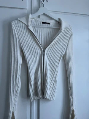 Vit ribbstickad hoodie med dragkedja - Säljer en vit ribbstickad hoodie från GINA TRICOT med dragkedja framtill. Tröjan har långa ärmar, huva och en figurnära passform. Perfekt för dig som gillar en enkel men snygg stil.