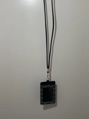 Svart mönstrad plånbok med snöre - Snygg svart plånbok med geometriskt mönster och genomskinlig ficka på framsidan. Plånboken har ett långt snöre så du kan bära den runt halsen. Perfekt för kort och ID. Stilren och praktisk accessoar för dig som vill ha koll på det viktigaste.