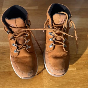 Bruna boots från Helly Hansen - Säljer ett par bruna vinterskor från Helly Hansen med grov sula och snörning. Skorna är i skinn och har metallhakar för snörningen samt HH-logga på sidan och under sulan. Perfekta för höst och vinter med sin robusta design och bekväma passform.