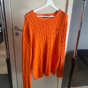 Orange kabelstickad tröja av Ralph Lauren - Säljer en orange kabelstickad tröja från Ralph Lauren med klassisk rund halsringning och broderad logga på bröstet. Tröjan har långa ärmar och ribbade muddar vid ärmslut och nederkant. Perfekt för dig som vill sticka ut med färg. Säljer den pågrund av att jag växt ur den.