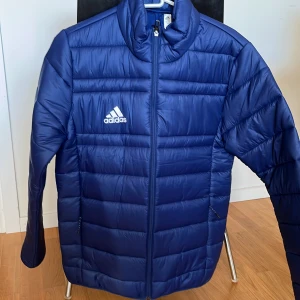 Blå pufferjacka från Adidas - Snygg blå pufferjacka från Adidas, i tunnare modell med klassisk logga på bröstet. Jackan har horisontella quiltade paneler, dragkedja framtill och två sidofickor med dragkedja. Perfekt för kyliga dagar och har en modern, sportig look. Jackan är aldrig använd, så i nyskick. 