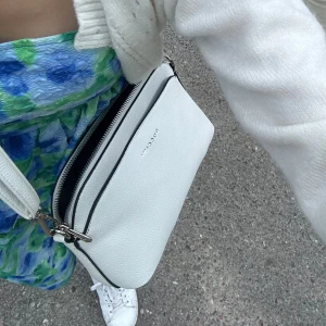 Vit axelväska från Puccini - Snygg vit axelväska från Puccini med stilren design och silverfärgade detaljer. Väskan är tillverkad i skinnimitation med struktur, har justerbar axelrem och flera fack med dragkedja. Perfekt storlek för mobil, plånbok och andra essentials.