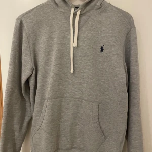 Grå hoodie från Polo Ralph Lauren - Snygg grå hoodie från Polo Ralph Lauren med klassisk blå logga på bröstet. Tröjan har huva med vit snörning, stor magficka och långa ärmar. 
