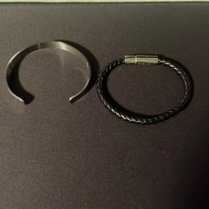 Två armband, Edblad & silverfärgat - Säljer två snygga armband! Ett svart flätat läderarmband från Edblad med magnetlås i silverfärg, och ett stilrent öppet armband i blankt silverfärgat metall. Perfekt för dig som gillar minimalistisk och modern stil. Skick på båda armbanden 9/10. Nypris Edblad armbandet 499kr. Säljer båda för 299!