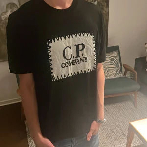Svart C.P. Company t-shirt  - Svart t-shirt från C.P. Company med ett stort vitt tryck på bröstet där märkesnamnet står i svart. Klassisk passform med rund hals och korta ärmar. Tillverkad i mjuk bomull som känns skön mot huden. Är storlek M men passar L