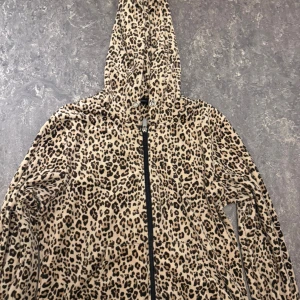 Leopardmönstrad teddyjacka med huva - Säljer en leopardmönstrad teddyjacka med huva och dragkedja framtill. Jackan är beige med svarta och bruna fläckar, har långa ärmar och är gjord i mjukt teddy-material. Perfekt för dig som gillar djurmönster och vill ha en cozy vibe.
