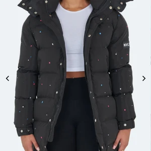 Svart lång pufferjacka från NVLTY - Svart lång pufferjacka från NVLTY med färgglada prickar i blått, rosa och vitt. Jackan har huva, dragkedja och knappar framtill samt elastiska muddar vid ärmslut. Perfekt för kalla dagar och har en cool streetstil-vibe.