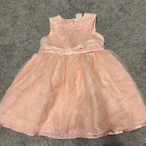 Ljusrosa spetsklänning från Lindex storlek 80 - Superfin ljusrosa kortklänning från Lindex med spetsdetaljer och ett brett satinband med rosett i midjan. Klänningen är ärmlös och har ett fluffigt kjolparti med lager av tyll och spets. Perfekt för speciella tillfällen och ger en söt look.