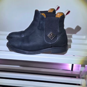 Svarta chelsea boots från Gant, Nyskick, Stl 38 - Svarta chelsea boots från Gant i skinn med klassisk rund tå och elastiska paneler på sidorna. Nyskick. Snygg detalj med Gant-logga på utsidan och dragflik med röd rand bak. Insidan är fodrad i brunt läder för extra komfort. ☆Skorna är nyimpregnerade för optimalt väderskydd och längre hållbarhet.☆
