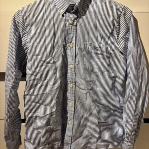 Blårandig skjorta från GANT - Snygg blårandig skjorta från GANT, modell The Banker Casual Fit. Skjortan har klassisk krage, bröstficka med broderad logga och vita knappar framtill. Tillverkad i mjuk bomull och har lång ärm. Perfekt för dig som gillar stilrena och fräscha plagg.