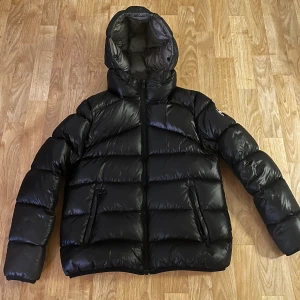 Svart Colmar pufferjacka med huva - Säljer en svart pufferjacka från Colmar med glansig finish och huva. Jackan har två dragkedjefickor fram, tydlig logga på ärmen och är fylld med naturligt dun. Perfekt för kalla dagar och riktigt snygg streetstil.