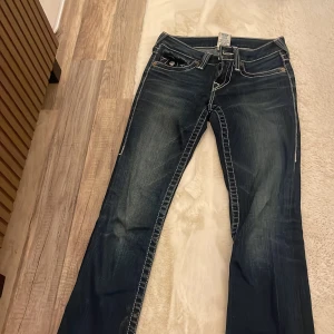 True Religion blå bootcut jeans - Säljer ett par blå jeans från True Religion med tydliga vita kontrastsömmar och klassiska fickdetaljer bak. Modellen har bootcut och låg midja, tillverkade i jeansmaterial med dragkedja och knapp framtill. Perfekt för dig som gillar street och statement.