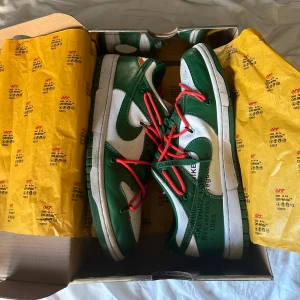 Nike Dunk Low Off-White Pine Green - Nike Dunk Low Off-White i färgen Pine Green och vitt, med unika röda extra snören och Off-White-typisk text på sidan. Skorna har låg profil, platt sula och är tillverkade i skinn. Kommer i äoriginal silverfärgad box med Off-White-papper. Orange snöre har en liten skada på högerskorn. 