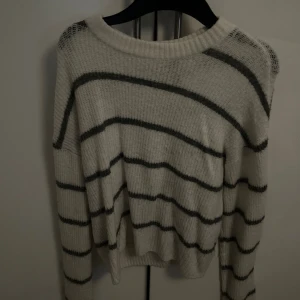 Vit stickad randig tröja - Mysig vit stickad tröja med svarta horisontella ränder. Tröjan har en rund halsringning och långa ärmar, perfekt för att hålla sig varm under kyliga dagar. Den har en loose passform och ribbad kant vid halsen och nederdelen.