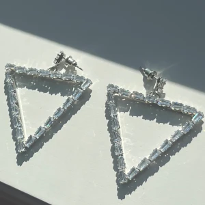 Glittriga triangelformade örhängen - Snygga örhängen i triangelform med glittrande stenar runt hela kanten. De är silvriga och har en modern, edgy vibe som verkligen sticker ut. Perfekta för dig som gillar att synas och vill ha något unikt till din stil.