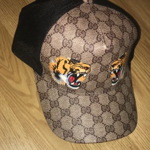  keps med tigertryck och GG-mönster - Snygg keps  med klassiskt GG-mönster i brunt och två coola tigertryck framtill. Baksidan är i svart mesh för extra ventilation. Märket syns tydligt på insidan och kepsen är tillverkad i polyester och bomull. Perfekt för dig som vill sticka ut med lyxig streetstyle.