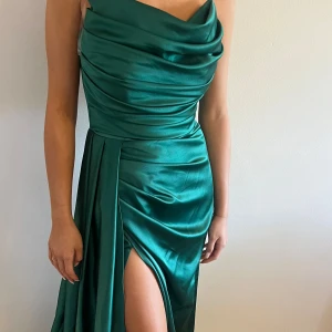 Grön långklänning i satin med slits - Kontakta mig om ni vill ha bättre bilder!      Elegant smaragdgrön långklänning/ balklänning i glansig satin med draperad överdel och djup slits framtill. Ser ut/ är en korsett upptill. Klänningen  har en figurnära passform och vackert fall, perfekt för att sticka ut. Axelbandslös design med svepande tygdetaljer längs sidan. Nypris var 3000 kronor!! Sk