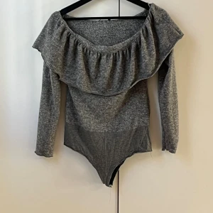 Grå volang body från Zara - Säljer en snygg grå body från Zara med bred volang runt axlarna och långärmad design. Toppen har offshoulder-stil och är tillverkad i mjukt syntetmaterial som sitter skönt mot kroppen. Perfekt för dig som gillar trendiga detaljer och vill sticka ut.