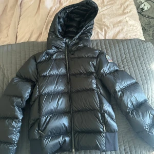 Svart dunjacka från Parajumpers - Säljer en svart dunjacka från Parajumpers med huva och snygga breda quiltade sektioner. Jackan har dragkedja framtill och två sidofickor med dragkedja. Märkespatch på ärmen och ribbade muddar vid nederkant. Perfekt för kalla dagar och riktigt stilren.