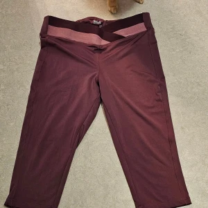 Vinröda träningsleggings från Crivit - Snygga vinröda träningsleggings från Crivit i storlek L. De har en bred, tvåfärgad midjeresår i vinrött och ljusrosa och är tillverkade i ett stretchigt syntetmaterial. Modellen är trekvarts-längd och passar perfekt för dig som gillar att röra på dig.