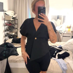 Svart omlottväst med guldknapp - Stilren svart väst med omlott-design och en snygg guldig knapp framtill. Vet ej vart den är ifrån, köpt på Plick. 