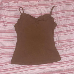 Brun ribbad linnetopp med volangkant - Säljer en brun linnetopp med smala axelband och volangkant längs halsringningen. Toppen är ribbad och har en lätt v-ringad form framtill. Perfekt för dig som gillar enkla men söta detaljer. Materialet känns mjukt och stretchigt.