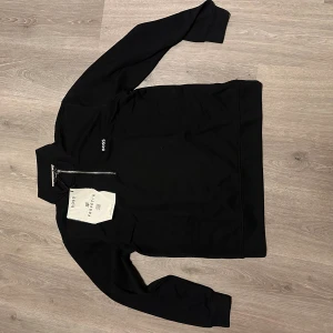 Svart half zip tröja från BOSS - Svart långärmad tröja från BOSS med stilren half zip-dragkedja och broderad logga på bröstet. Tillverkad i mjuk bomull och polyesterblandning, perfekt för dig som gillar minimalistisk och clean stil. Hög krage och normal passform.