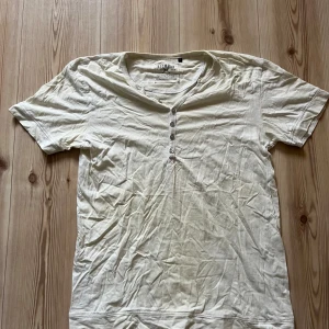 Vit t-shirt med knappar från Redwood strl M - Vit t-shirt från Redwood i storlek M. Modellen har korta ärmar, rundad hals och fyra knappar framtill som ger en chill och avslappnad vibe. Tillverkad i mjuk bomull som känns skön mot huden. Perfekt för dig som gillar enkel och stilren design.