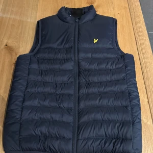 Mörkblå dunväst från Lyle & Scott - Sprillans ny nygg mörkblå dunväst från Lyle & Scott med gul logga på bröstet. Västen har quiltad design, hög krage och dragkedja framtill. Perfekt för lager-på-lager och enkel att matcha med hoodies eller tröjor.