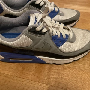 Nike Air Max 90 i grått och blått - Nike Air Max 90 sneakers i grått, vitt, svart och blått. Klassisk siluett med mesh och mockadetaljer, vit snörning och synlig Air Max-dämpning i sulan. Perfekta för dig som gillar streetstyle och sportig vibe.