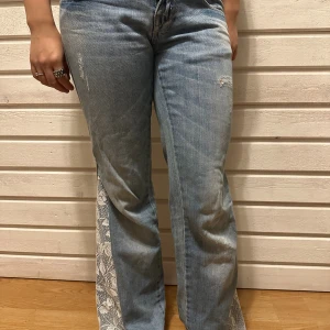 Blå bootcut jeans med spetsdetalj - Unika ljusblå jeans med bootcut-modell och breda spetsinslag i vitt längs bensluten. Jeansen har klassisk femficksdesign och subtila slitningar framtill. Spetsen har blommigt mönster som ger en cool och personlig touch till looken. Sytt själv. Innerbenslängd  75cm, midjemått 39cm