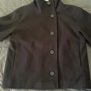 Svart kappa med hög krage från RYVLS - Stilren svart kappa från RYVLS med hög krage och raka linjer. Jackan har stora svarta knappar framtill och diskreta fickor i sidorna. Materialet är mjukt och känns som ull, perfekt för kyliga dagar. Passar dig som gillar minimalistisk och clean look.