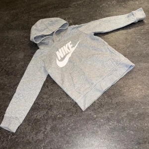 Grå hoodie från Nike med logga - Grå Nike hoodie med stor vit logga på bröstet. Tröjan har huva, lång ärm och en klassisk känguruficka framtill. Mjuk insida och ribbade muddar vid ärmslut och nederkant. Perfekt för chill eller träning.
