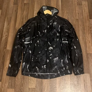 Svart camo vindjacka från SOC - Svart vindjacka med grått camo-mönster från SOC. Jackan har huva, hel dragkedja och reflekterande detaljer. Tillverkad i lätt syntetmaterial, perfekt för blåsiga dagar. Långärmad och med meshpanel på insidan för extra ventilation.