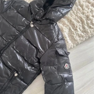 Svart pufferjacka med huva - Svart pufferjacka med glansig yta, huva och dragkedja framtill. Jackan har två sidofickor med dragkedja och en ficka på ärmen med logga. Perfekt för kalla dagar och har en riktigt snygg streetstil.