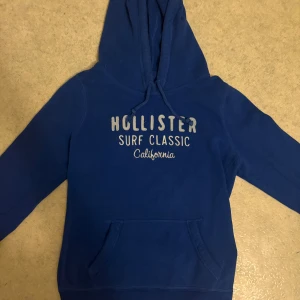Blå hoodie från Hollister - Snygg marinblå hoodie från Hollister med tryck 'Surf Classic California' på bröstet. Tröjan har huva med snörning, stor magficka och långa ärmar. Mjuk insida i fleece och klassisk passform, perfekt för chill dagar. Står storlek L passar storlek S inte några specifika defekter utan att den är använd. Varan är inte urtvättad.