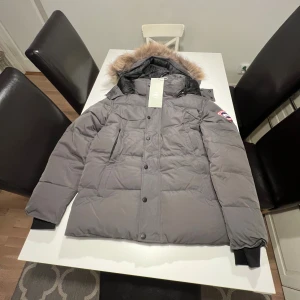 Grå dunjacka Canada Goose XS - Säljer en grå dunjacka från Canada Goose i storlek XS men den sitter som en lite större S. Jackan har en fluffig huva med päls, ribbade muddar och klassisk logga på ärmen. Fylld med dun och har knappar framtill. Perfekt för kalla vinterdagar och har en clean, stilren look.